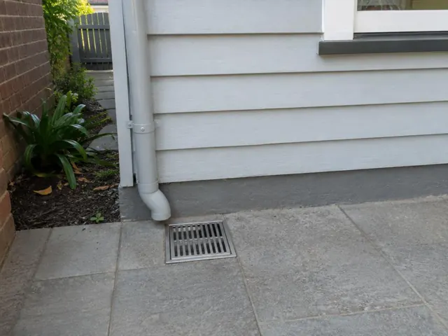 Outdoor drain grate beside a downpipe on grey paving next to a house wall in Neutral Bay